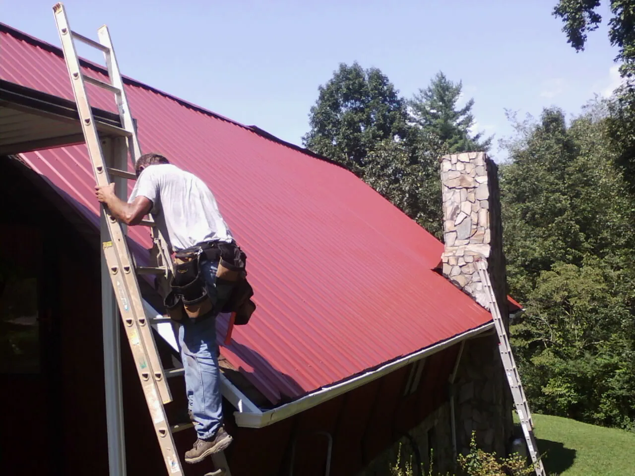 Roofer on a ladder beside a red metal roof and stone chimney — Soffit & Fascia Repair by Roofing Brothers in Cohocton